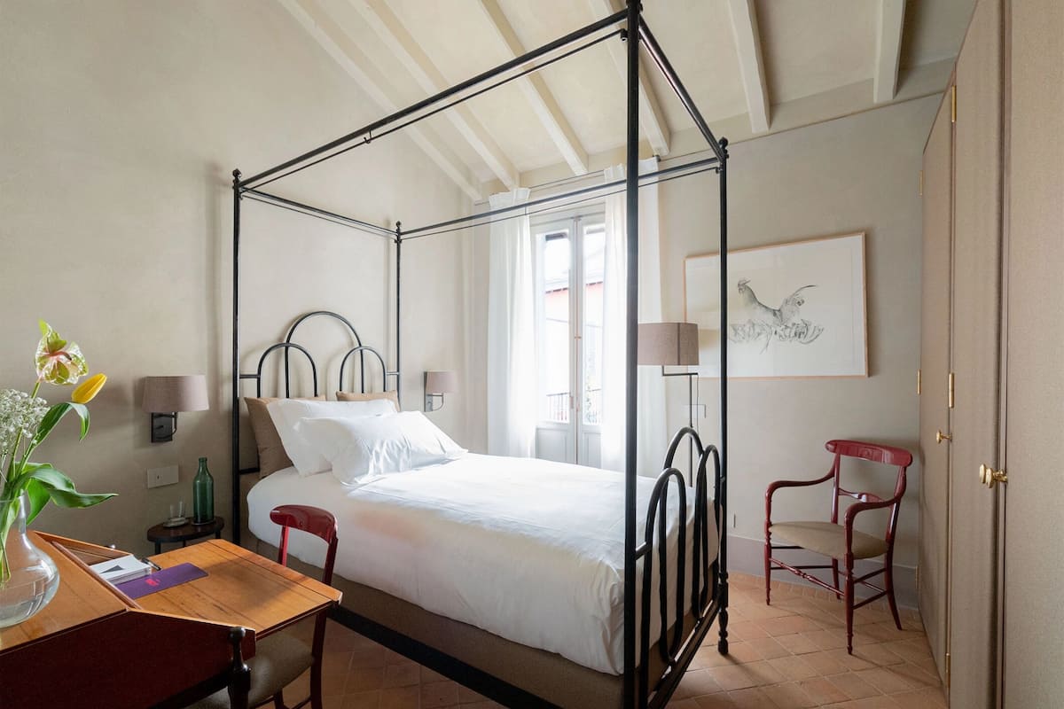 A four-poster bed with a wooden headboard, a bedside table with a lamp, a chair, and a vase with flowers.