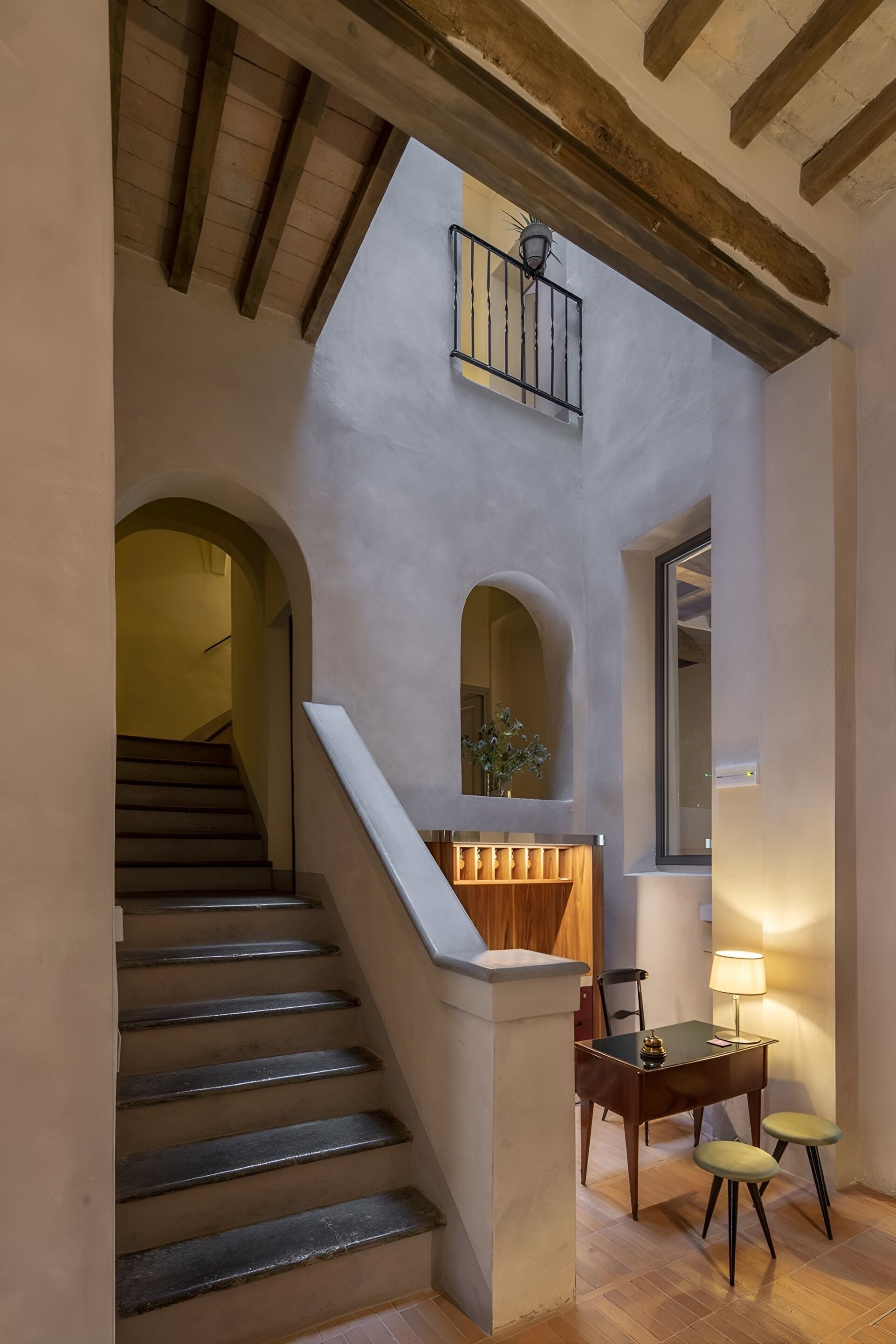 A staircase with a wooden handrail leading to a landing with a table and two chairs, a small balcony, and an arched doorway.