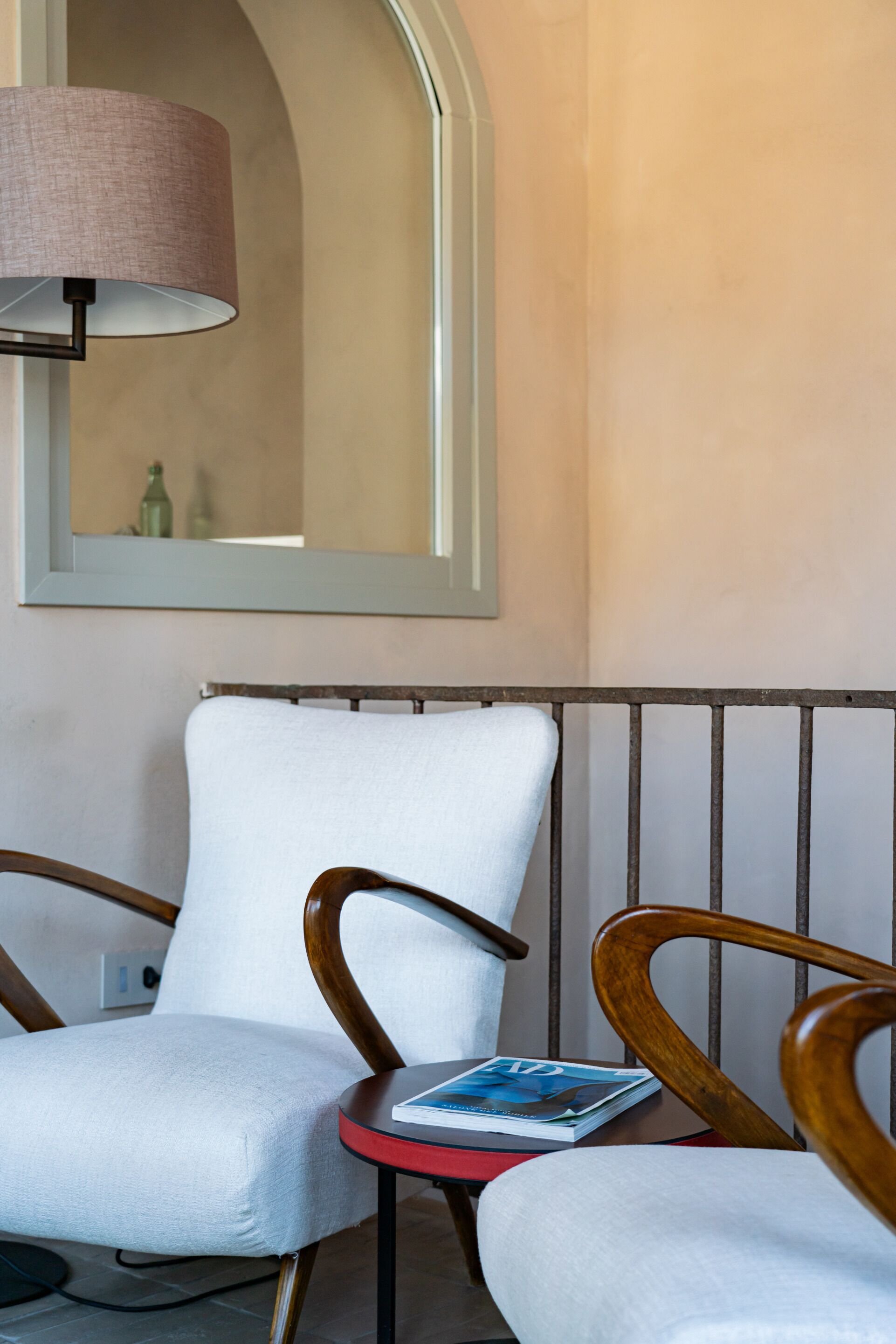 Une pièce avec un fauteuil blanc, une chaise en bois et une petite table sur laquelle se trouve un magazine. Une lampe est fixée au mur, et un miroir reflète une partie de la pièce.