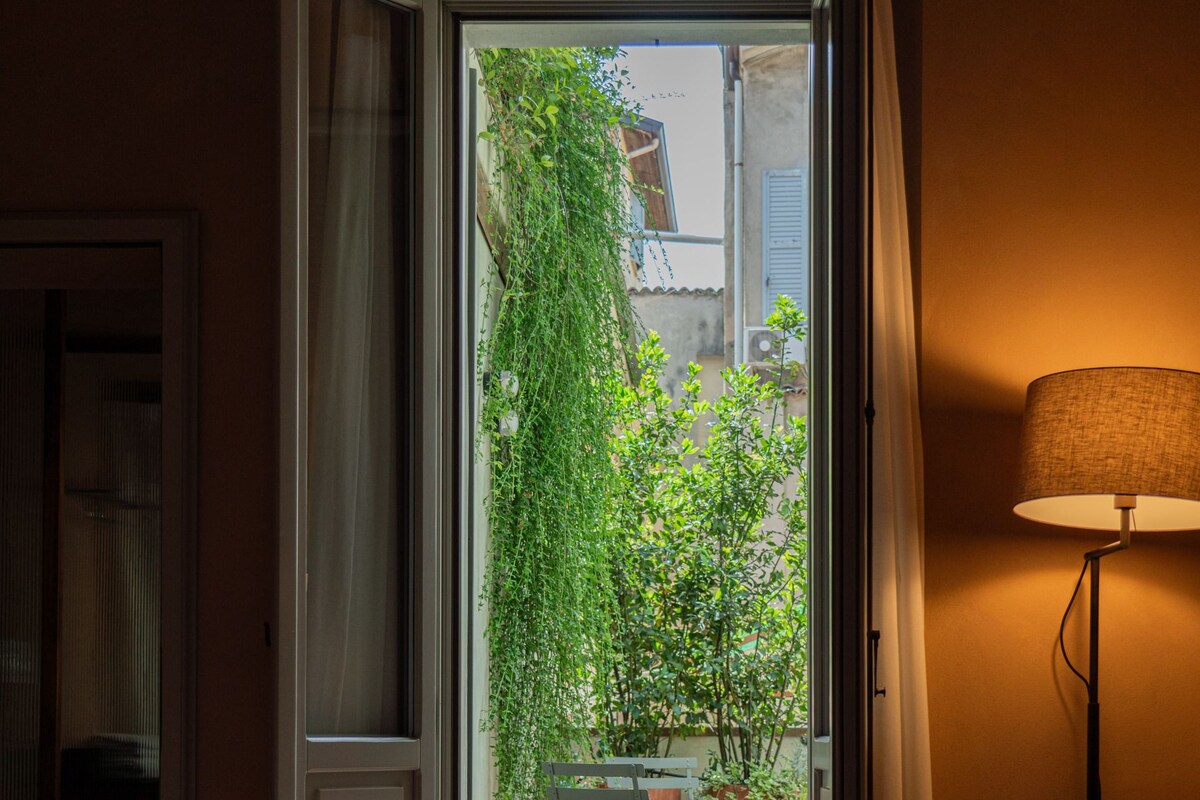 Habitación de hotel con una cama, una silla, una mesa y una puerta abierta que da a un espacio exterior con vistas a zonas verdes y un edificio.