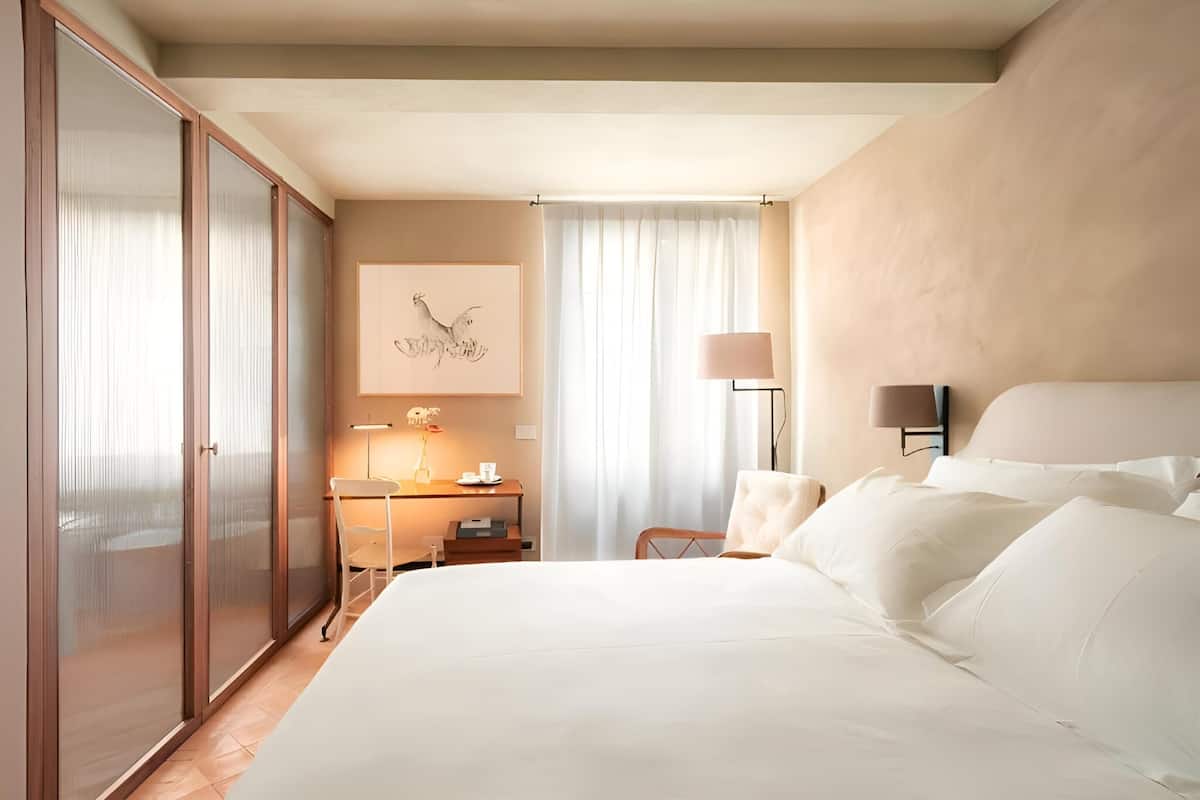 A hotel room with a large bed, a desk with a lamp, a chair, and a wardrobe.