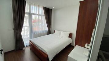 A hotel room with a large bed, wooden flooring, a white bedside table, and a window with curtains.