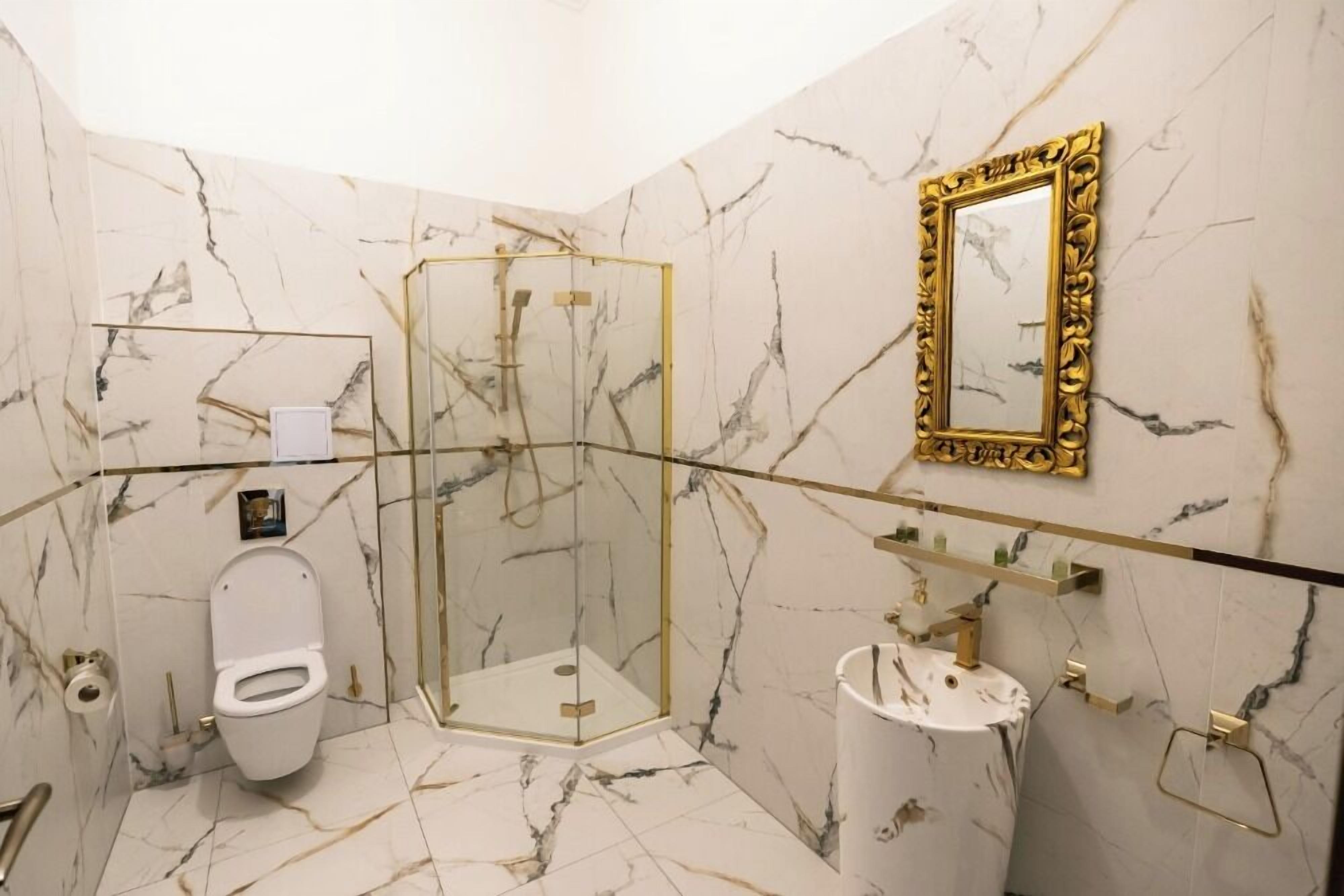 Un baño con piso de mármol, ducha envuelta en vidrio, inodoro, lavamanos y espejo con marco dorado.