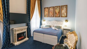Een slaapkamer met een open haard, een bed met een blauwe sprei, een nachtkastje met een lamp en drie ingelijste foto’s aan de muur.