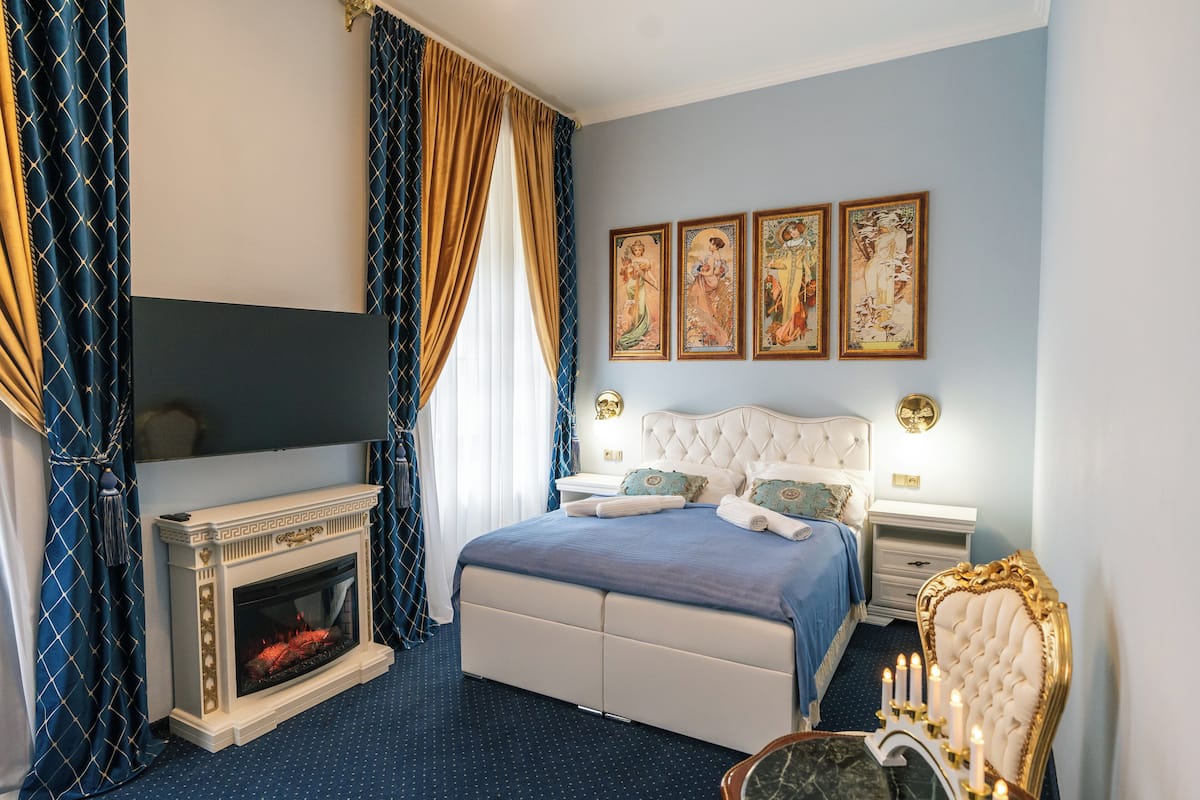 A bedroom with a fireplace, a bed with a blue bedspread, a nightstand with a lamp, and three framed pictures on the wall.