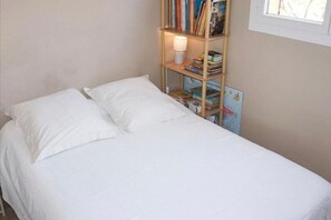 A single bed with white bedding, a bedside lamp, and a bookshelf with books and a picture frame.