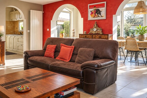 A living room with a brown leather sofa, red cushions, a wooden coffee table with a bowl of colorful balls, and a dining area with wicker chairs.