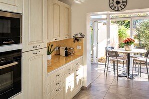 A kitchen with light wood cabinets, a black oven, and a tiled backsplash. There is a dining area with a table and chairs, and a view of the outdoors through a glass door.