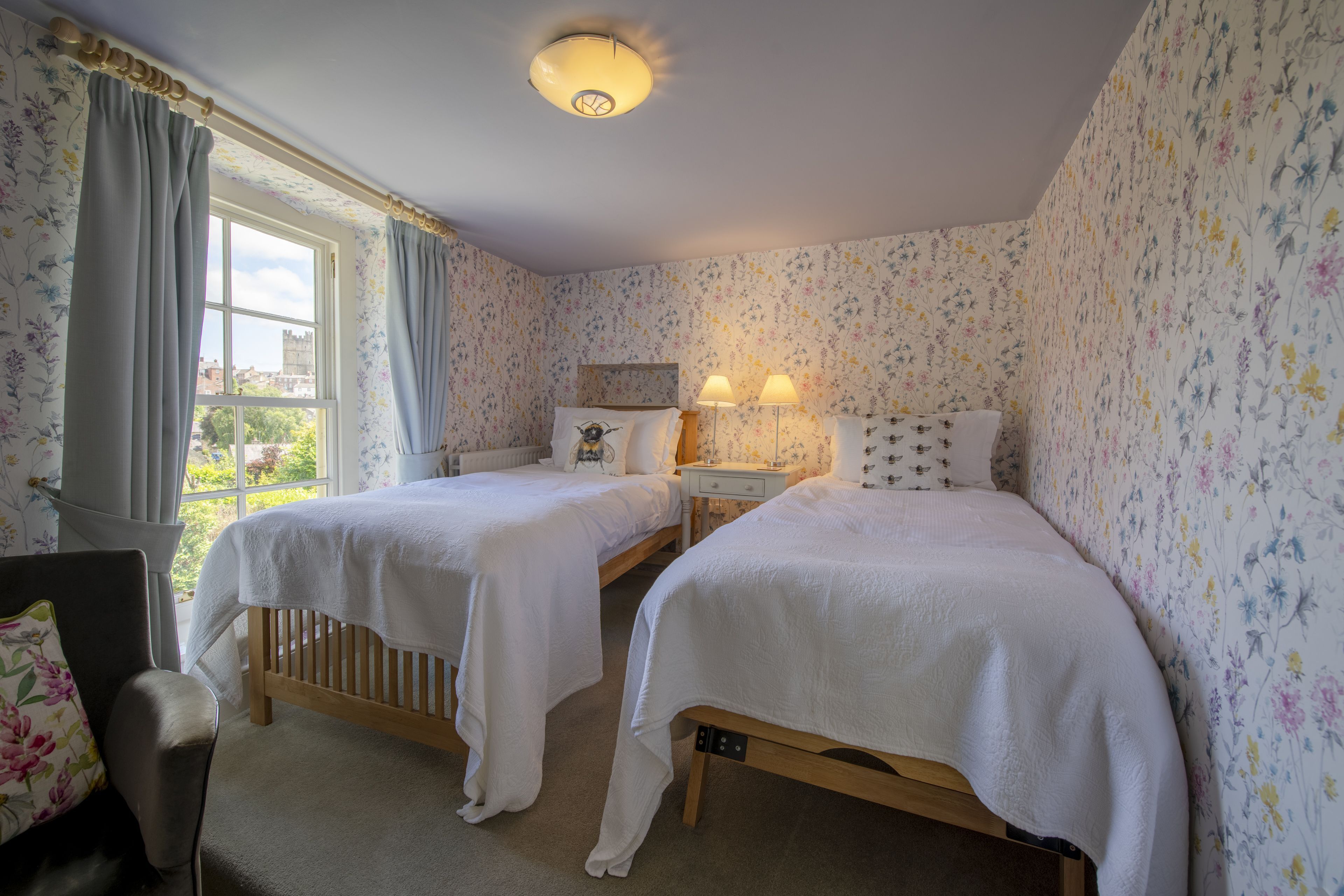 A room with two single beds, a nightstand with lamps, floral wallpaper, and a view of greenery outside.