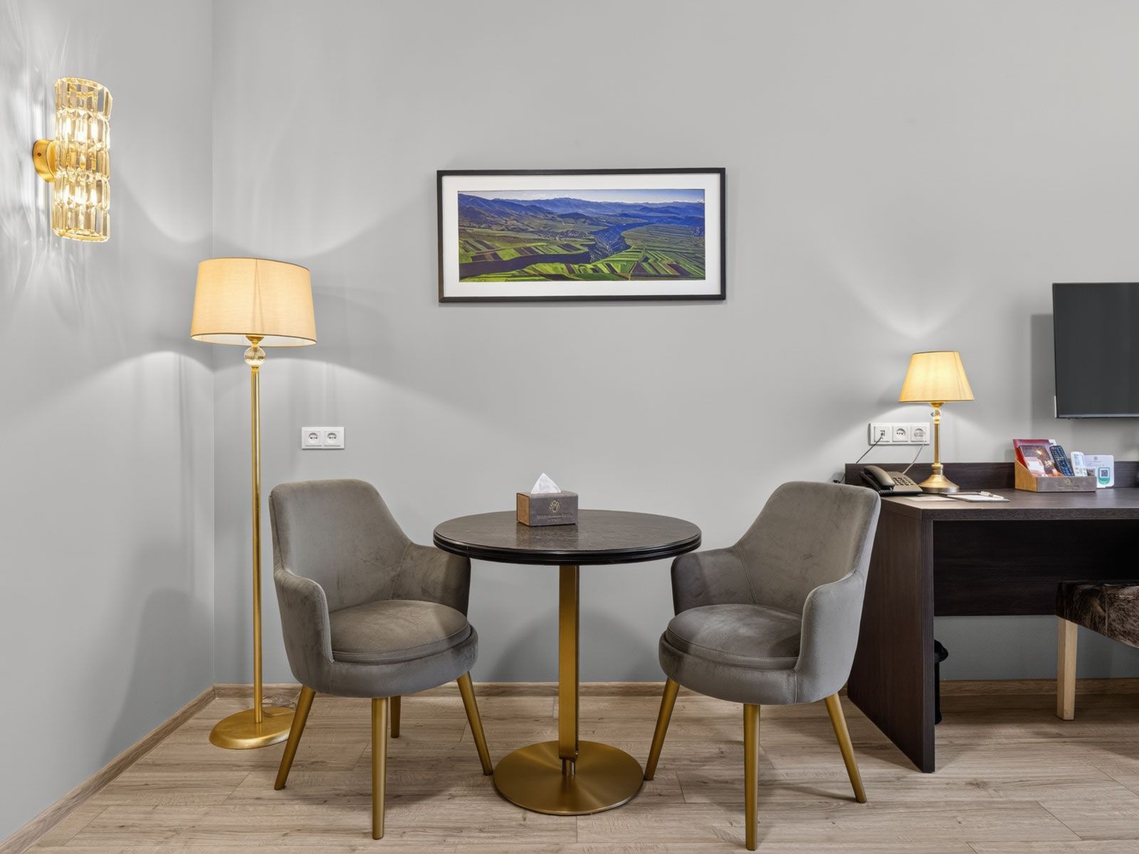 Quarto de hotel com uma mesa redonda, duas cadeiras, uma escrivaninha com um computador, um abajur, uma pintura de paisagem emoldurada e uma luminária fixada na parede.