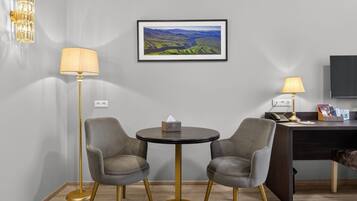 Quarto de hotel com uma mesa redonda, duas cadeiras, uma escrivaninha com um computador, um abajur, uma pintura de paisagem emoldurada e uma luminária fixada na parede.