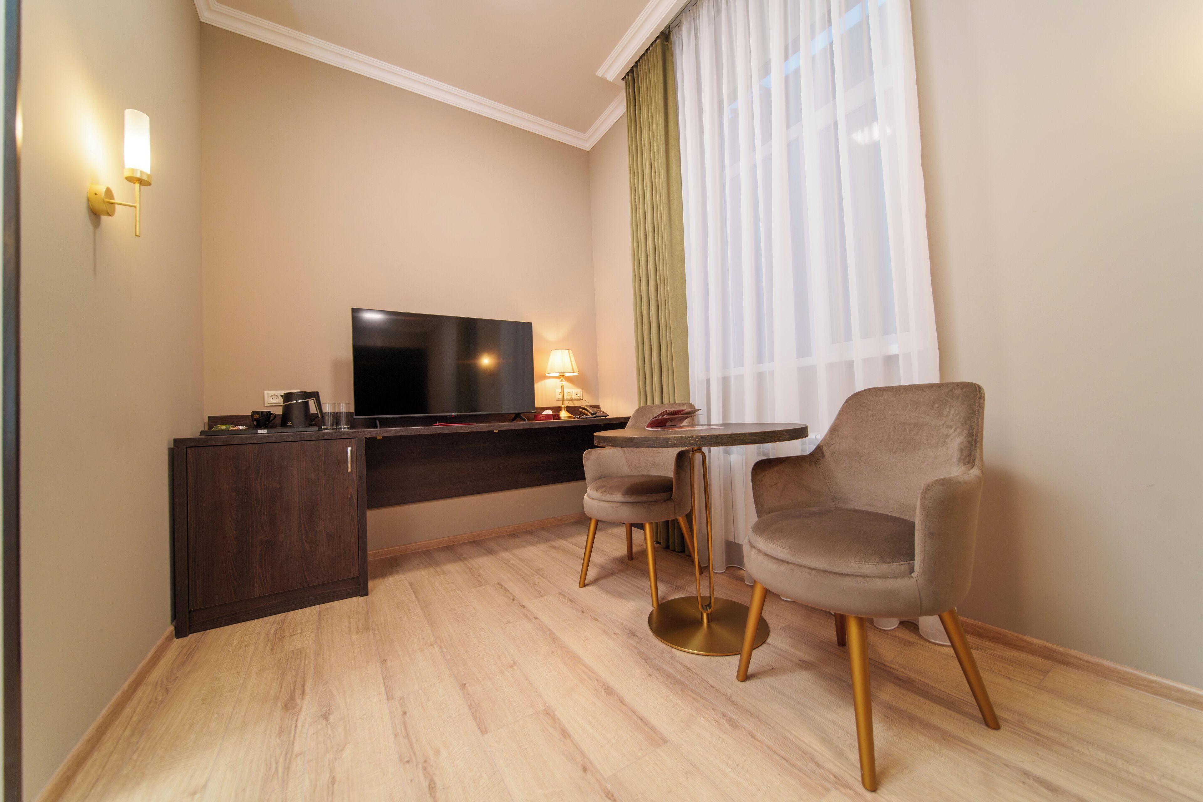A modern hotel room with a flat-screen TV, a small dining table, two chairs, a dark wooden desk, and a lamp.