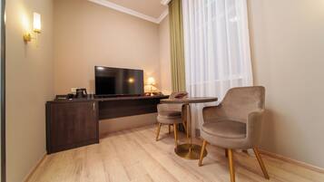 A modern hotel room with a flat-screen TV, a small dining table, two chairs, a dark wooden desk, and a lamp.