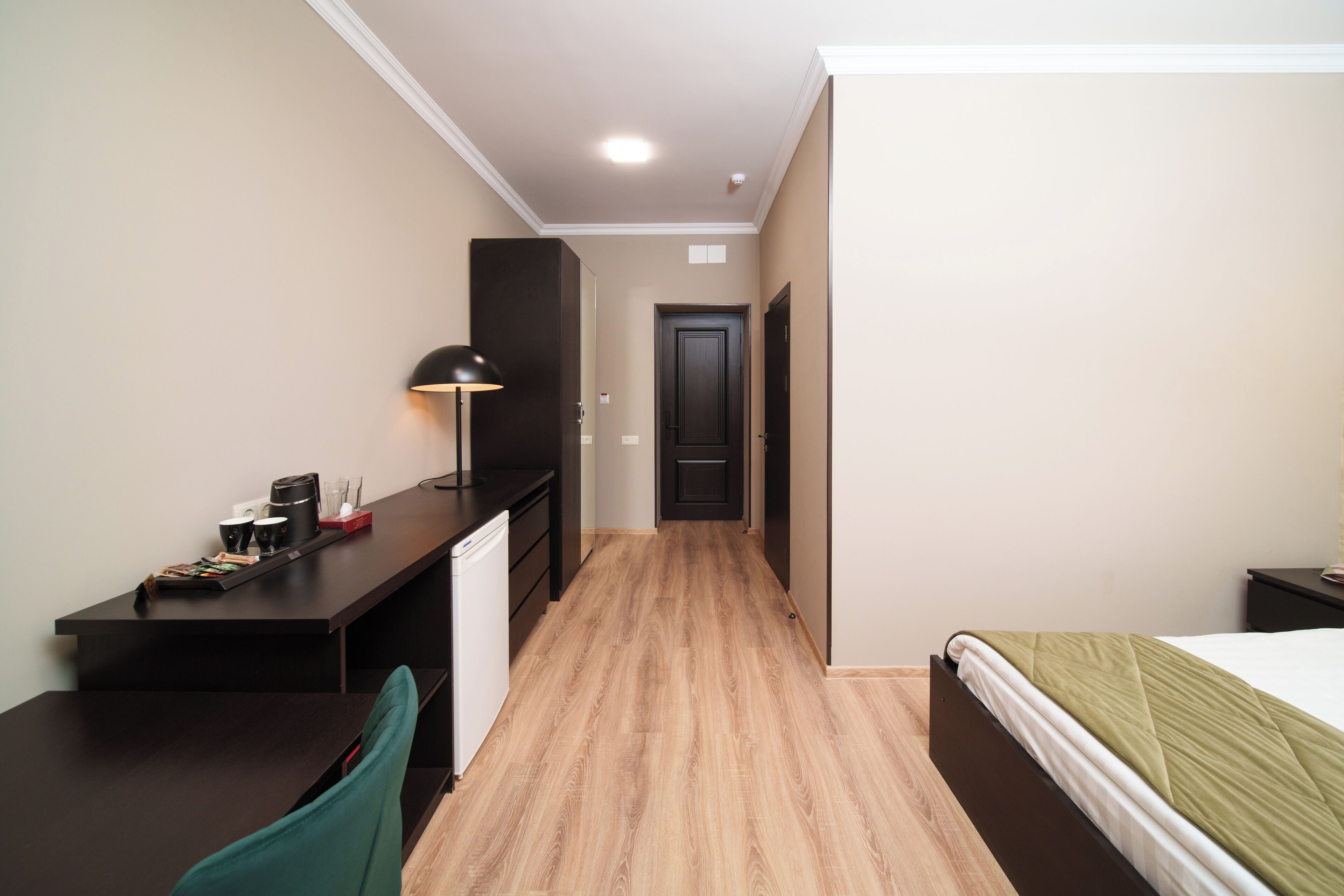 A hotel room with a bed, a desk, a chair, a lamp, and a door.
