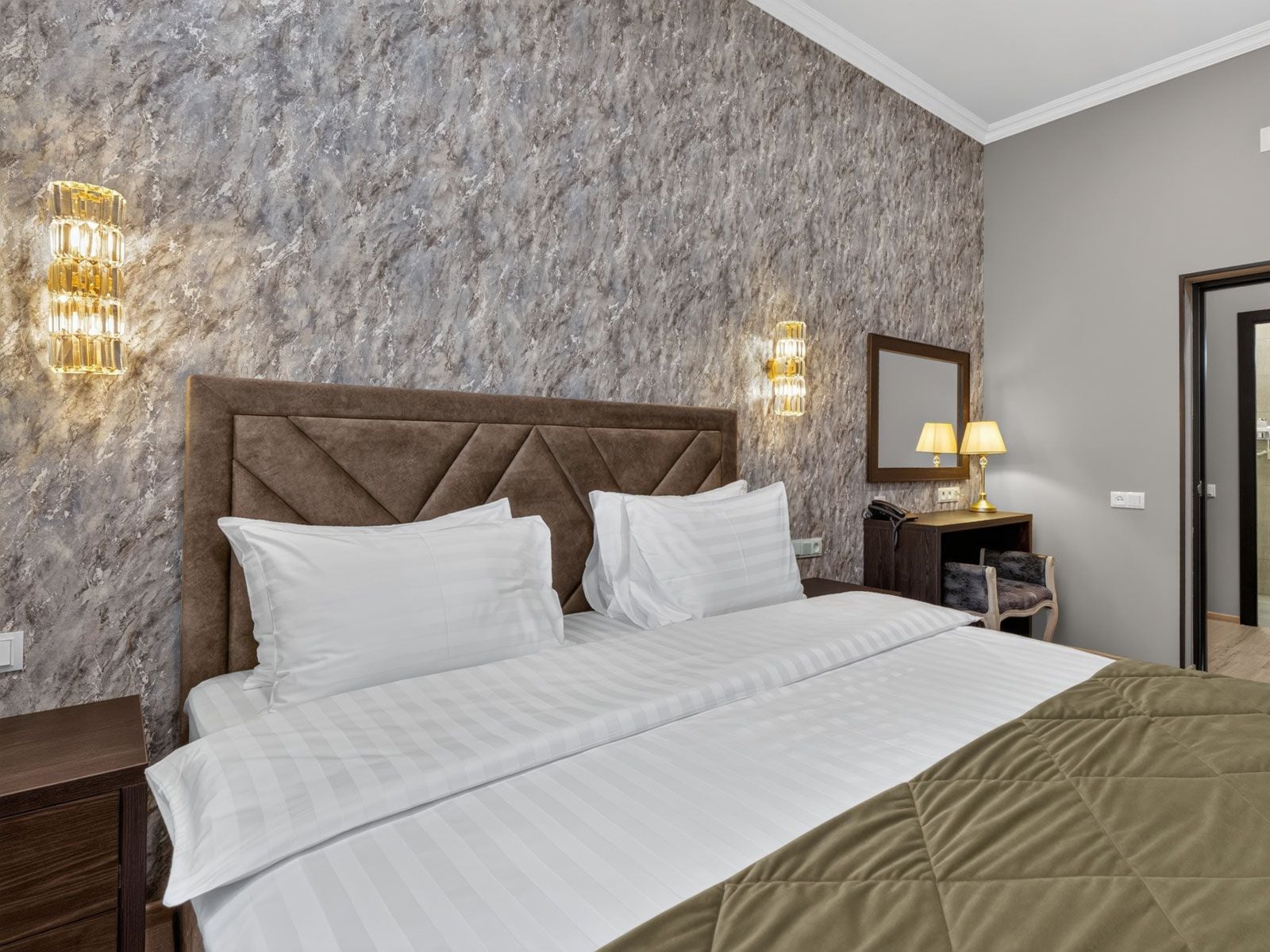Habitación de hotel con pared de diseño de mármol, cabecera capitonada, cama con ropa blanca, dos lámparas de noche y mesita de noche con lámpara.