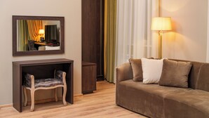 A modern hotel room with a wooden console table, a cushioned armchair, a brown sofa with two pillows, a floor lamp, and a large mirror reflecting the room.