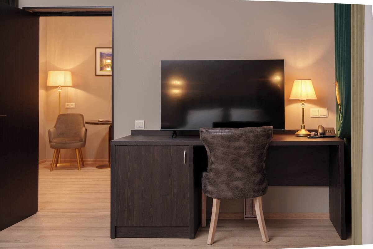 A hotel room with a dark wood desk, a leather chair, a flat-screen TV, two lamps, and a green curtain.