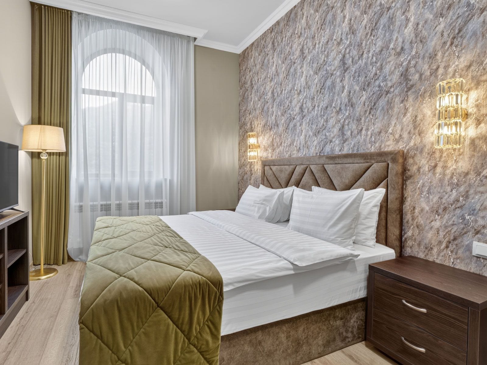A hotel room with a bed, a bedside table, a lamp, and a wall-mounted light fixture.