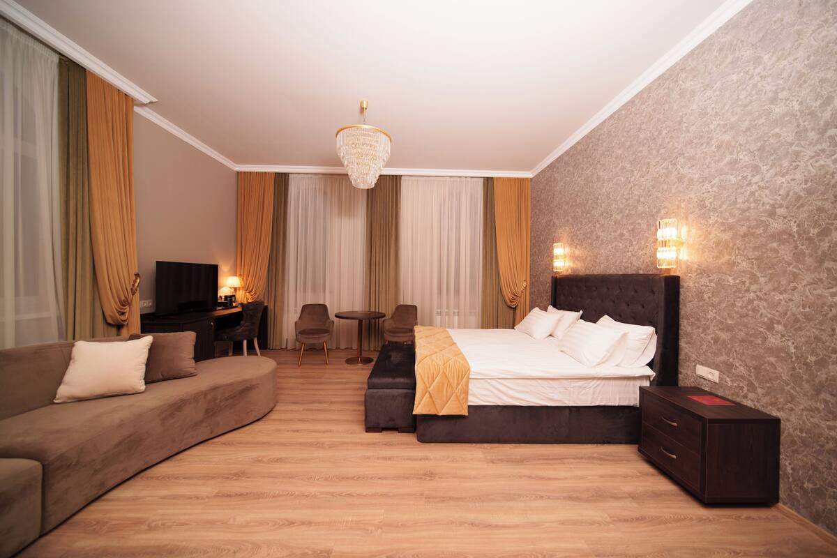 A hotel room with a large bed, a sofa, a desk with a chair, a TV, and a chandelier.