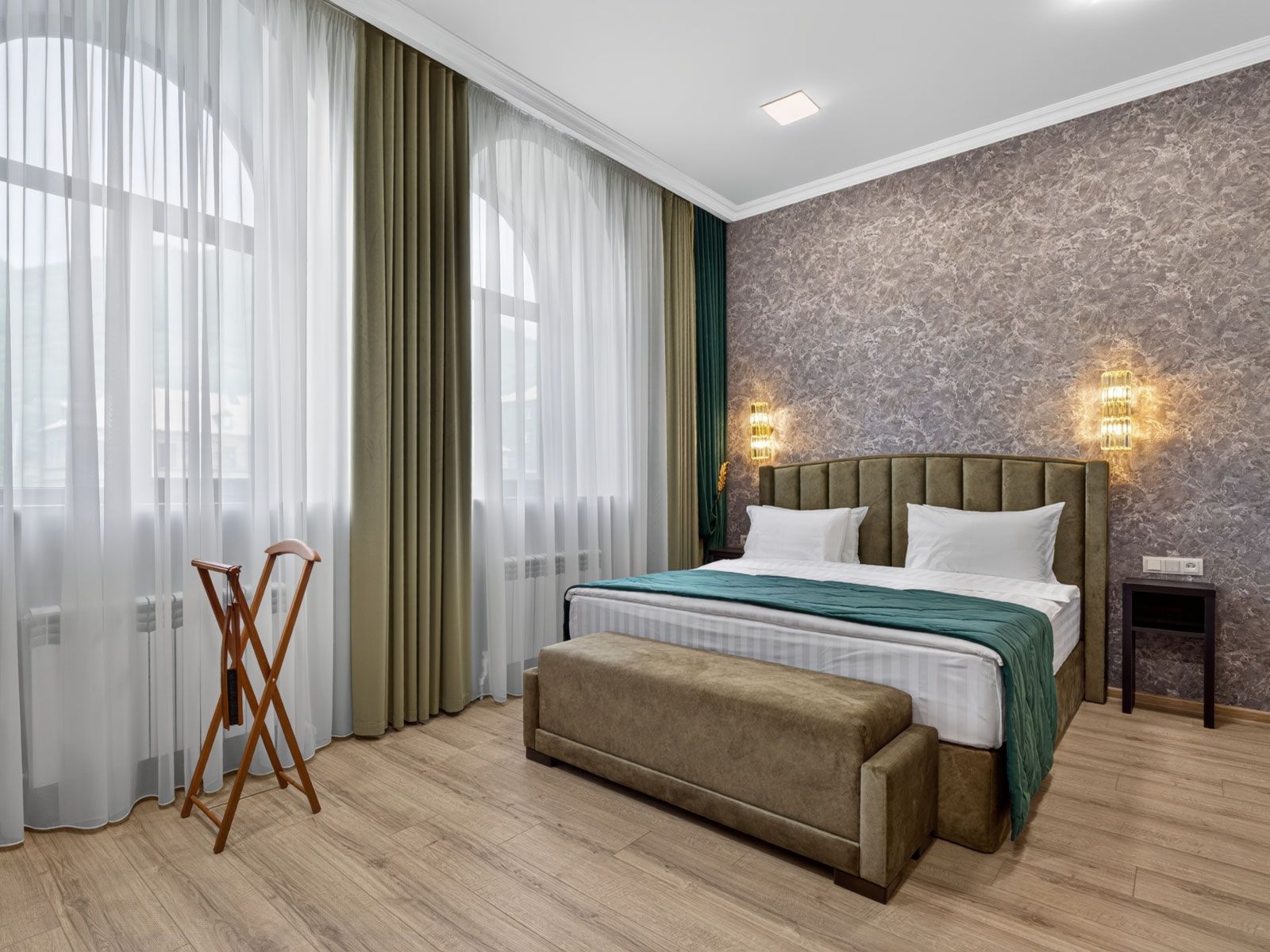 Quarto com cama, um banquinho, luminária de chão e janela com cortinas.