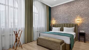 Quarto com cama, um banquinho, luminária de chão e janela com cortinas.