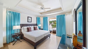 A spacious bedroom with a large bed, two bedside tables, a ceiling fan, and a balcony with blue curtains. - Smugglers Cove 6 (4 bed): Beachfront condo (Paynes Bay)