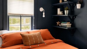 Un dormitorio con cama, ventana con persianas, estante en la pared con libros y adornos, y un reloj de pared.