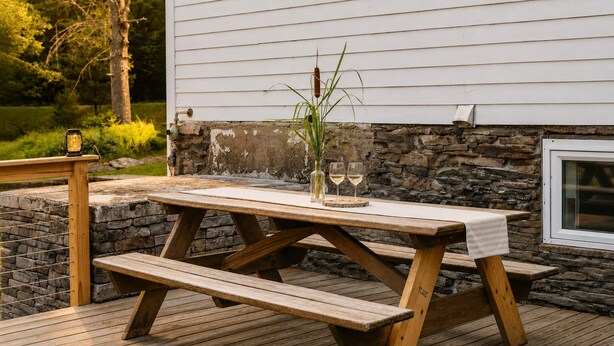 Een houten picknicktafel met bankjes op een houten terras, voor een stenen muur en een wit gebouw.