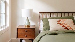 Een slaapkamer met een houten nachtkastje, een lamp, een bed met groene beddengoed, en een raam.