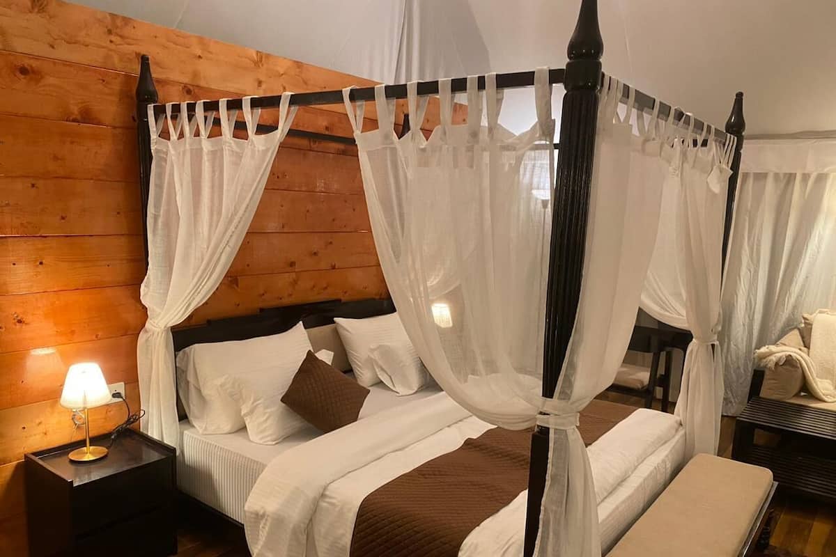 Una cama con dosel, ropa de cama blanca y una manta marrón, una lámpara de noche y un cabecero de madera.
