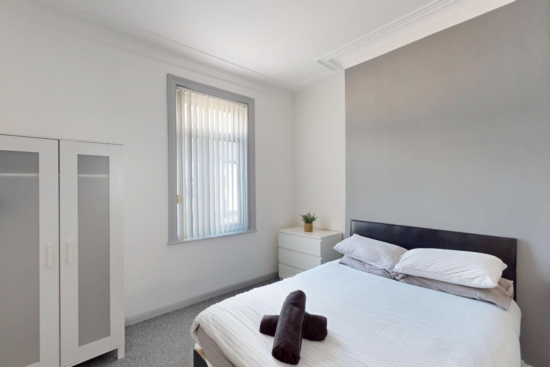 A bedroom with a bed, a wardrobe, a bedside table, and a window with blinds.