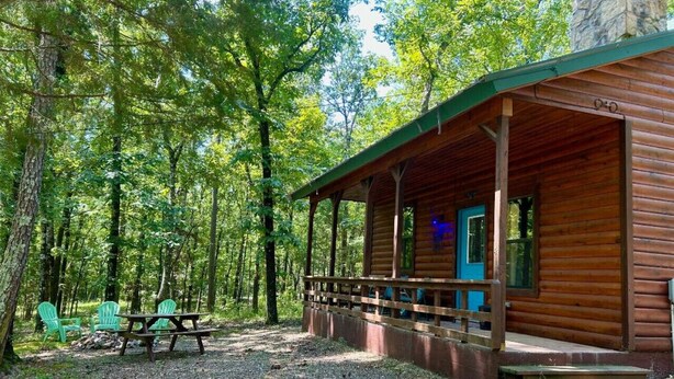 A wooden cabin with a porch, picnic table, and green chairs in a forested area. - Lost In The Woods Cabin | Hochatown Cabin with Hot Tub, Fire Pit, Grill & Pets (Hochatown)