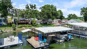 A waterfront property with docked boats, a covered deck area, and residential houses in the background. - Pet-Friendly Lakefront Home in a Quiet Cove w/ Netflix, Outdoor Seating, Boat Slip (Climax Springs)