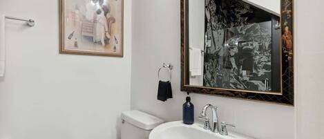 A bathroom with a white sink, a toilet, a framed picture on the wall, and a mirror with a black frame.