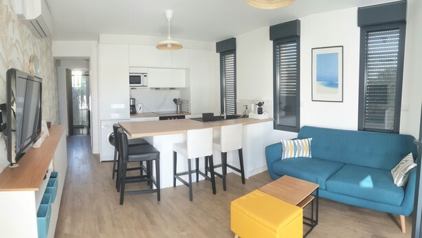 Eine moderne Küche mit weißen Schränken, einer zentralen Kochinsel mit Barhocker, einem blauen Sofa mit gemustertem Kissen und einem gelben Hocker.