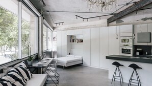 Un intérieur moderne et minimaliste, doté d’une grande fenêtre, d’un coin repas, d’un lit et d’une kitchenette.