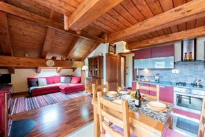 A cozy loft-style room with a wooden ceiling, a dining area with a table and chairs, a kitchen with a stove and cabinets, and a red sofa with cushions.