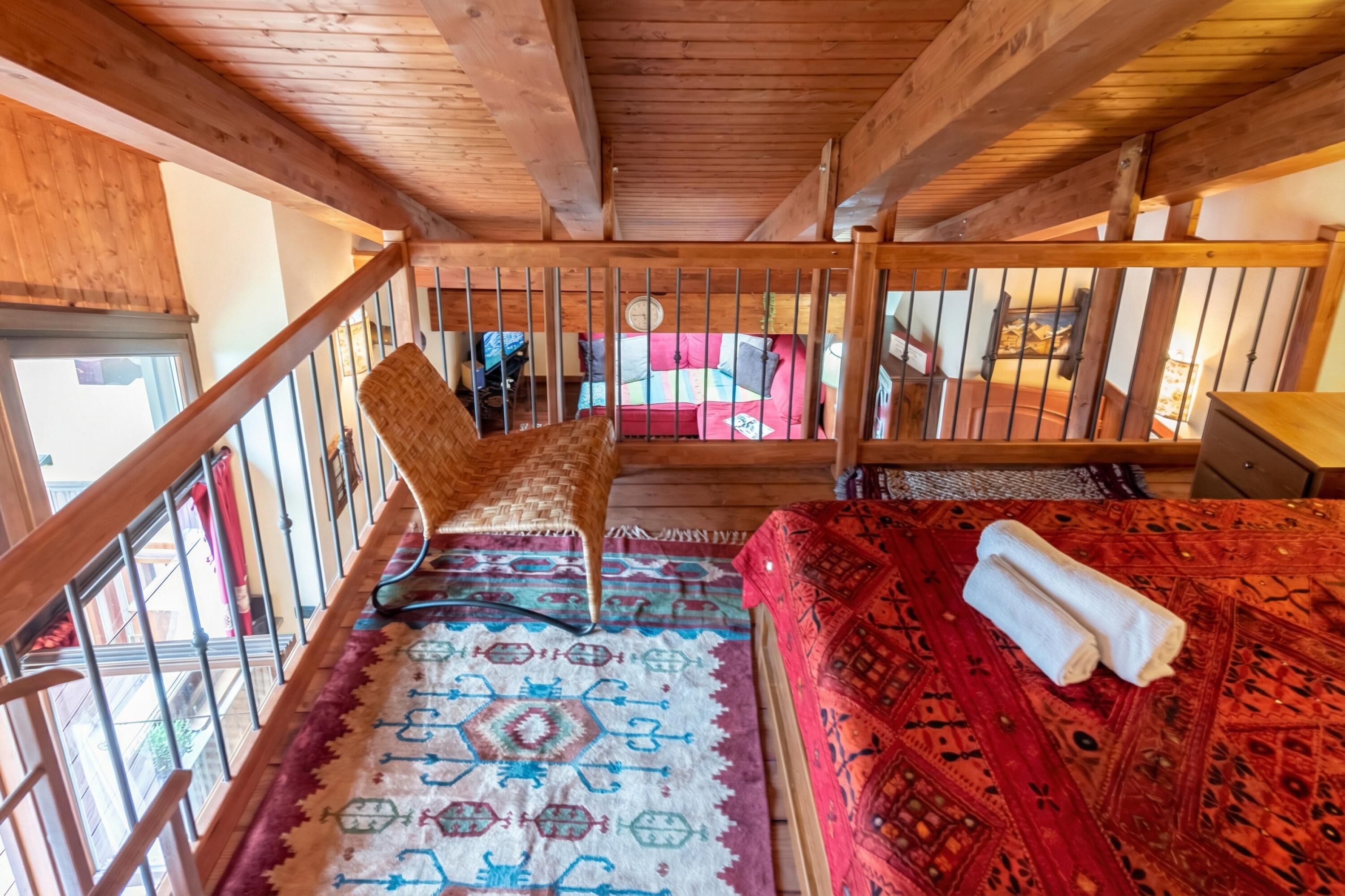 A wooden balcony with a woven chair, a patterned rug, and a bed with a red and black patterned bedspread.