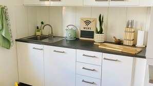 Una cocina moderna con armarios blancos, encimera negra y un hervidor verde. Se observa una tabla de cortar de madera, una planta y un cartel con un símbolo de WiFi.