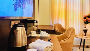 Habitación de hotel con televisor, cafetera, mesita con bandeja, una silla y una pequeña mesa con un jarrón de flores.