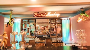 Una zona de bar con un letrero de neón que dice “Wanted Monkey”, estantes con botellas y una barra con vasos y una caja registradora.