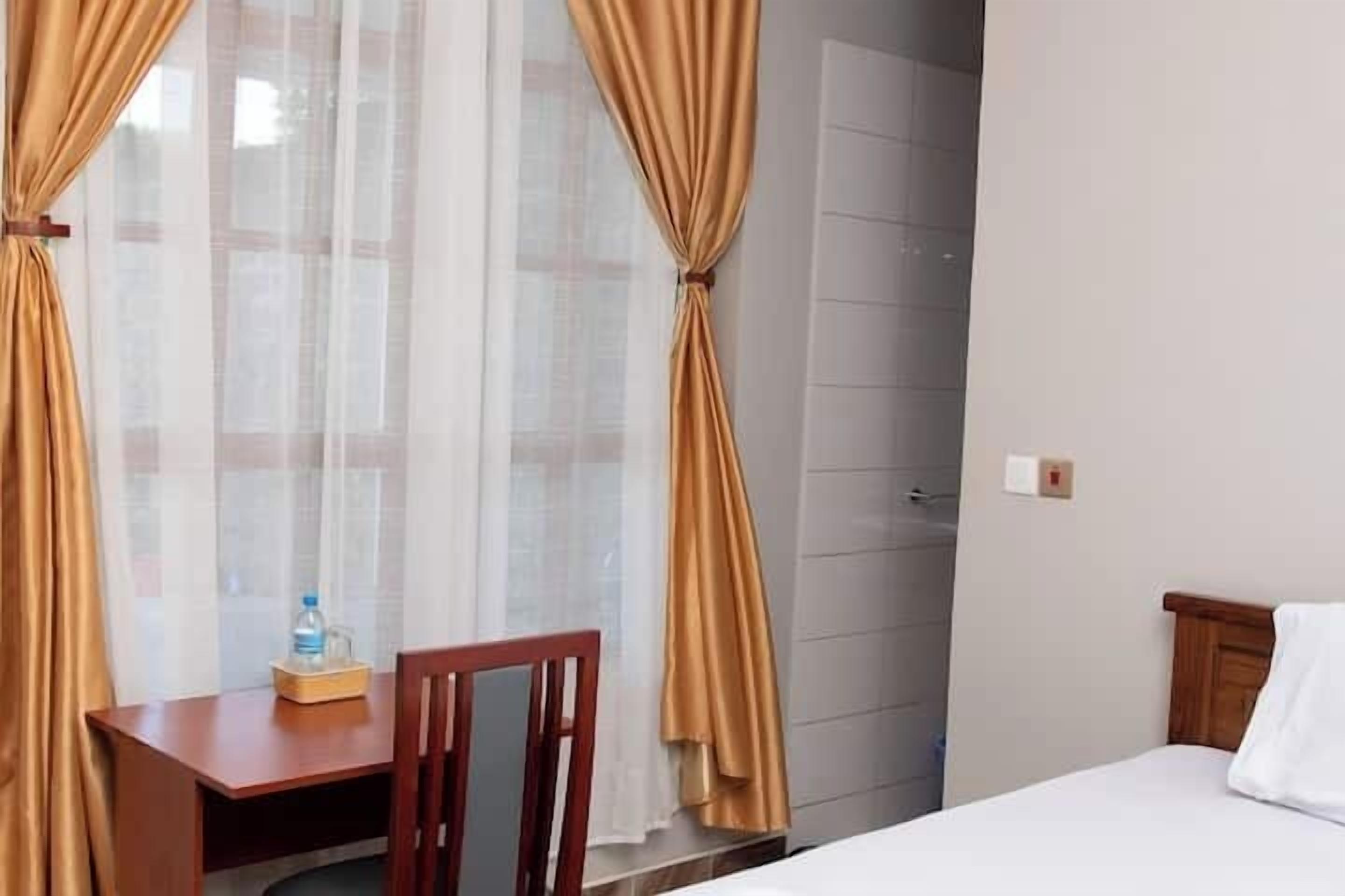 A hotel room with a bed, a wooden desk with a chair, a bottle of water, and curtains.
