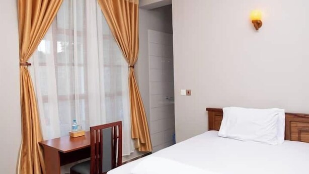A hotel room with a bed, a desk, and a chair. There is a window with curtains, a wall-mounted light, and a door. - Simba Villa (Arusha)