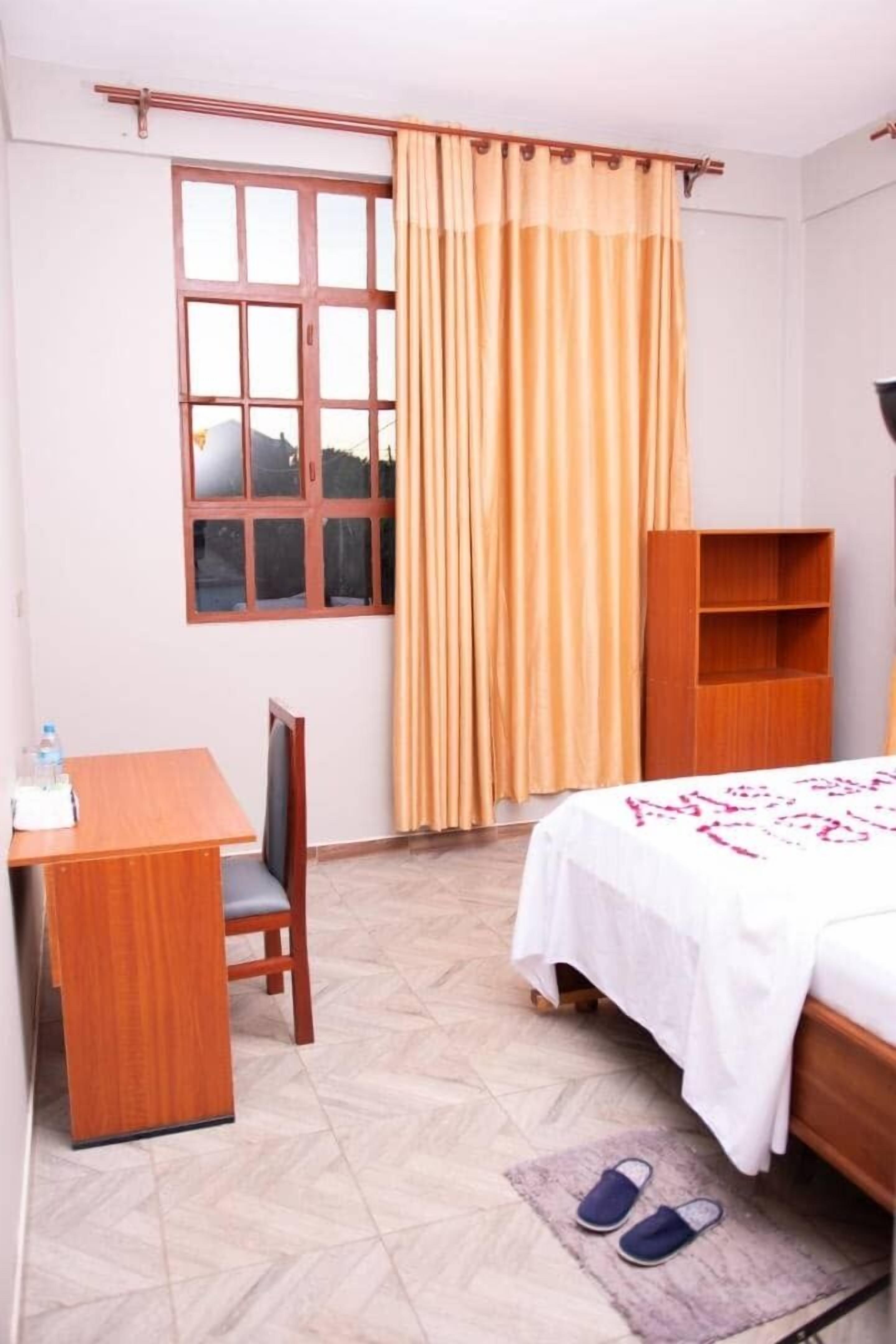 A hotel room with a bed, a desk, a chair, a wooden wardrobe, and a window with curtains.
