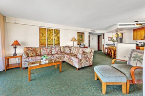 A living room with a sofa, a coffee table, and a bench. There is a kitchen area with wooden cabinets and a dining table.