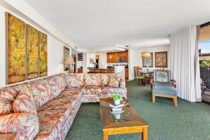 A spacious living room with a large sectional sofa, a coffee table with books and a plant, and a view of the kitchen and dining area through an open doorway.