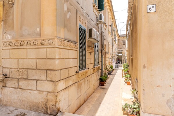 A narrow street with buildings on either side, featuring a tiled pavement and potted plants along the sidewalk.