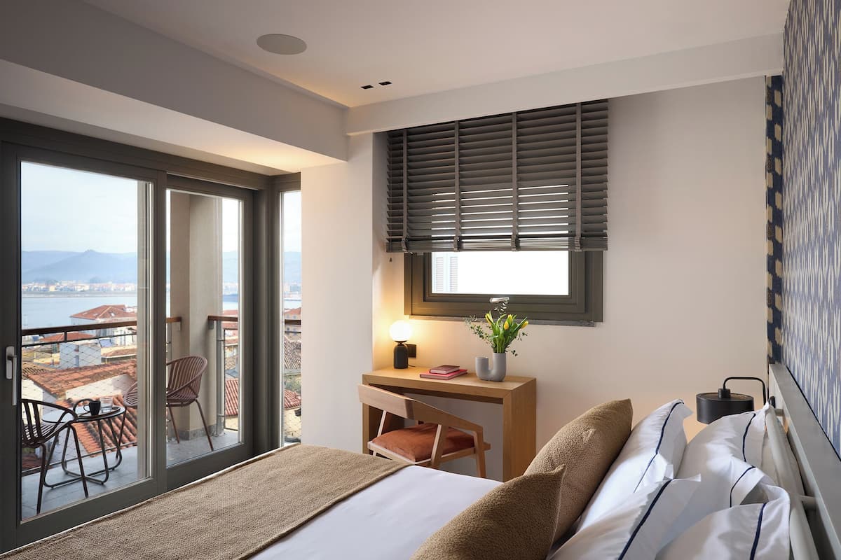 A hotel room with a bed, a desk, a chair, a window with blinds, a view of the city, and a balcony with chairs and a table.