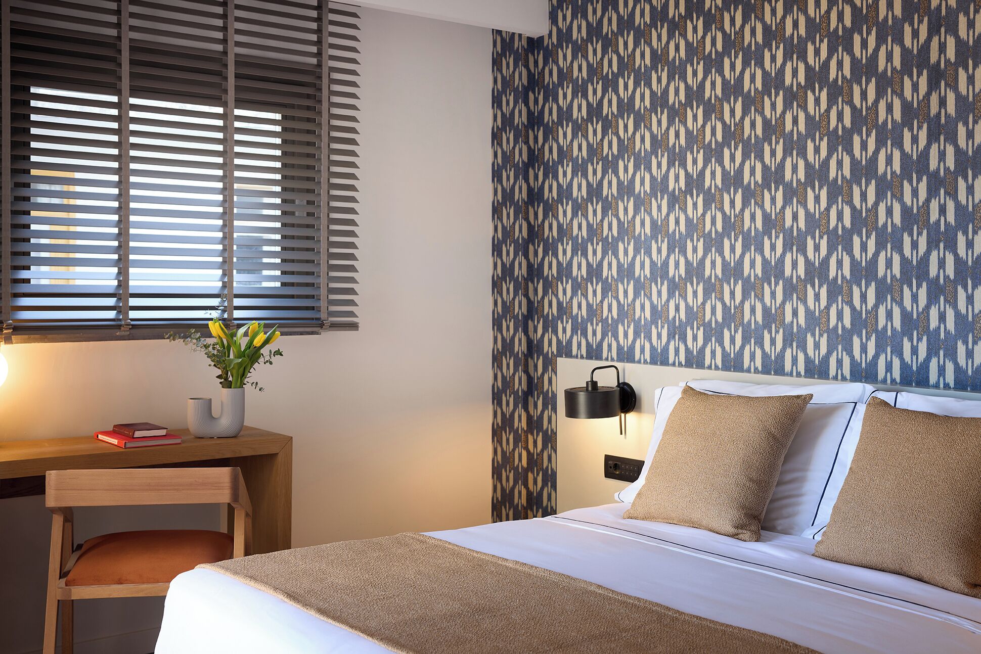 A bedroom with a bed, a desk, a lamp, and a patterned wall.