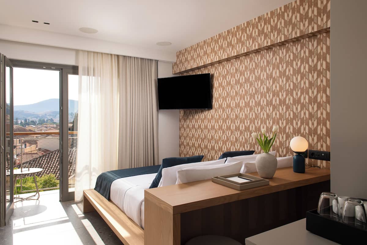A hotel room with a bed, a desk with a lamp, a TV, and a view of the city through a large window.